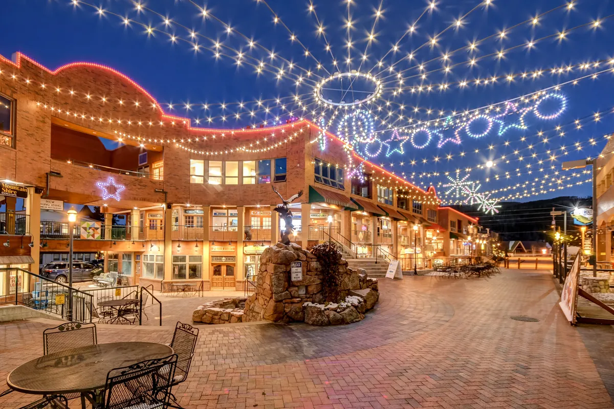 A lively downtown scene with decorative lights strung above, a bubbling stone fountain, and charming storefronts, conveying a festive and vibrant atmosphere. The image suggests socializing, shopping, and dining in a prime location.