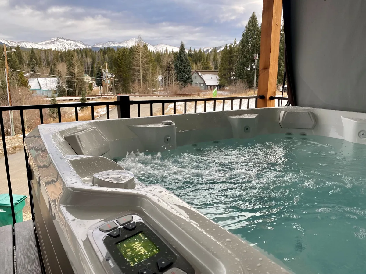 A tranquil exterior shot of a private hot tub set against a backdrop of mountains and surrounding trees. The scene suggests relaxation and luxurious comfort after a day of adventure, with steam rising from the hot tub and the golden glow of sunset in the air.