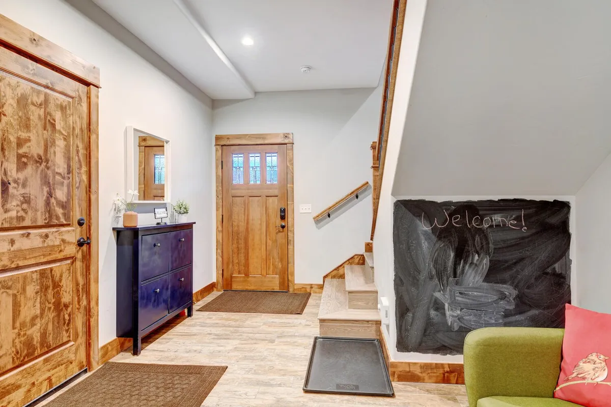 A welcoming entryway with a wooden front door, chalkboard wall, green couch, and mirror. The design is inviting and practical, giving the sense of a functional and homey transition space perfect for unloading adventure gear after a day outdoors.