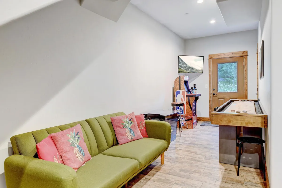 A casual, fun-filled lounge with a green sofa, colorful pillows, a shuffleboard table, and a wall-mounted TV. The setting is ideal for relaxed social gatherings, movie nights, and lively game sessions.
