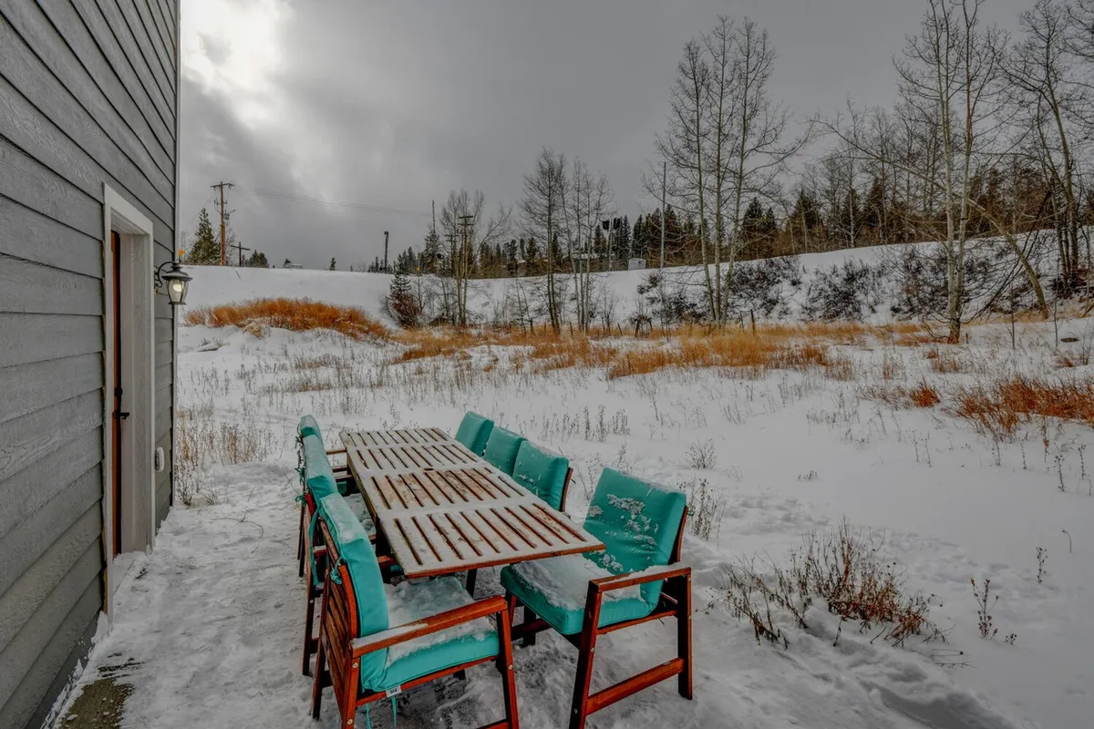 An outdoor dining area featuring a wooden table and turquoise chairs sits on a snow-covered patio, bordered by bare trees. The serene winter landscape extends into open space, suggesting easy access to outdoor adventures and walks.