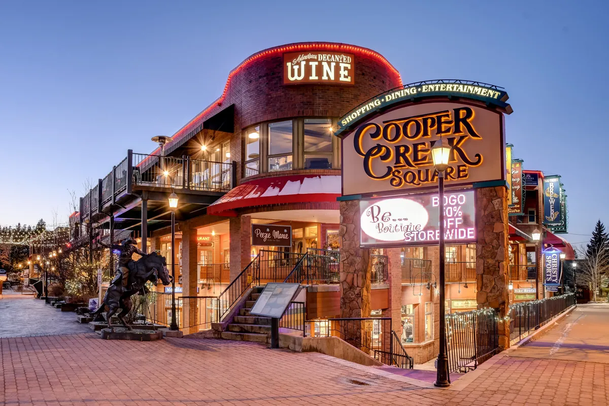 A lively scene of Cooper Creek Square, featuring its distinctive sign, warmly lit storefronts, outdoor seating, and a whimsical horse sculpture. Shoppers and diners gather in the welcoming plaza, evoking the vibrant heart of Winter Park's downtown.