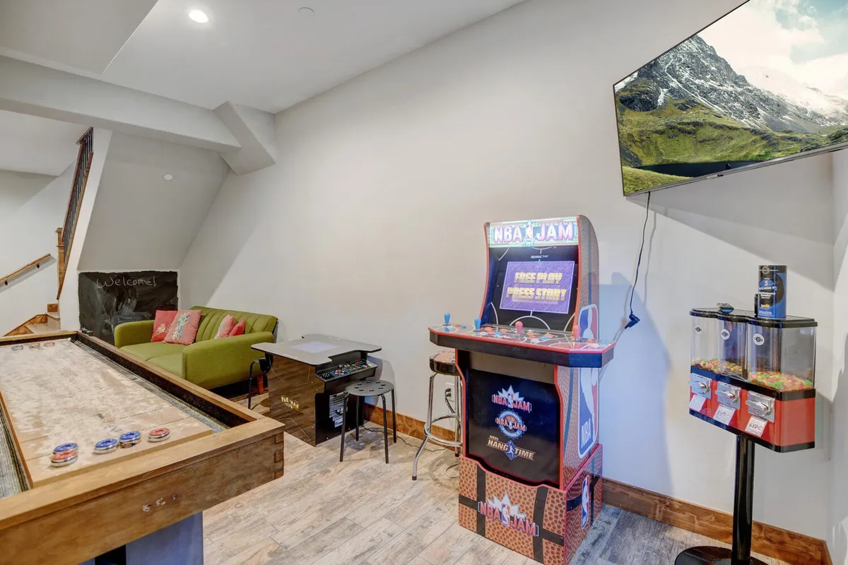 A lively game room featuring an NBA Jam arcade machine, shuffleboard table, green sofa with cushions, a TV displaying a scenic mountain view, and a candy dispenser. The scene is fun and relaxed, with visible opportunities for group play and social interaction.